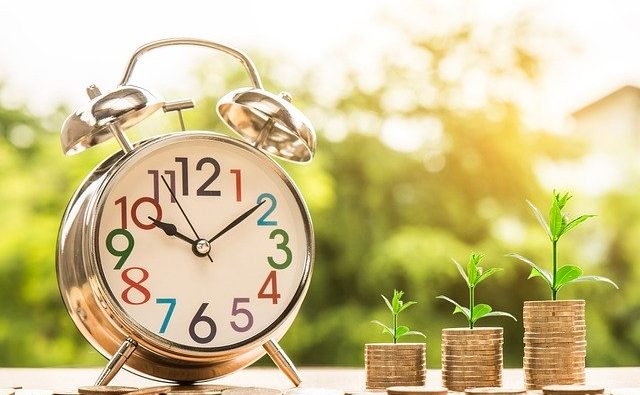 A silver alarm clock with multicolored numbers sits on a wooden surface beside three stacks of coins, each topped with small green plants. The background is blurred greenery, suggesting growth and prosperity over time. A silver alarm clock with multicolored numbers sits on a wooden surface beside three stacks of coins, each topped with small green plants. The background is blurred greenery, suggesting growth and prosperity over time.