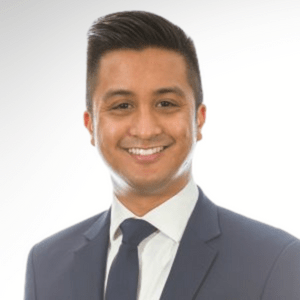 A man with short, dark hair is smiling while wearing a navy blue suit, a white dress shirt, and a navy blue tie. He is standing against a gradient white and light gray background.