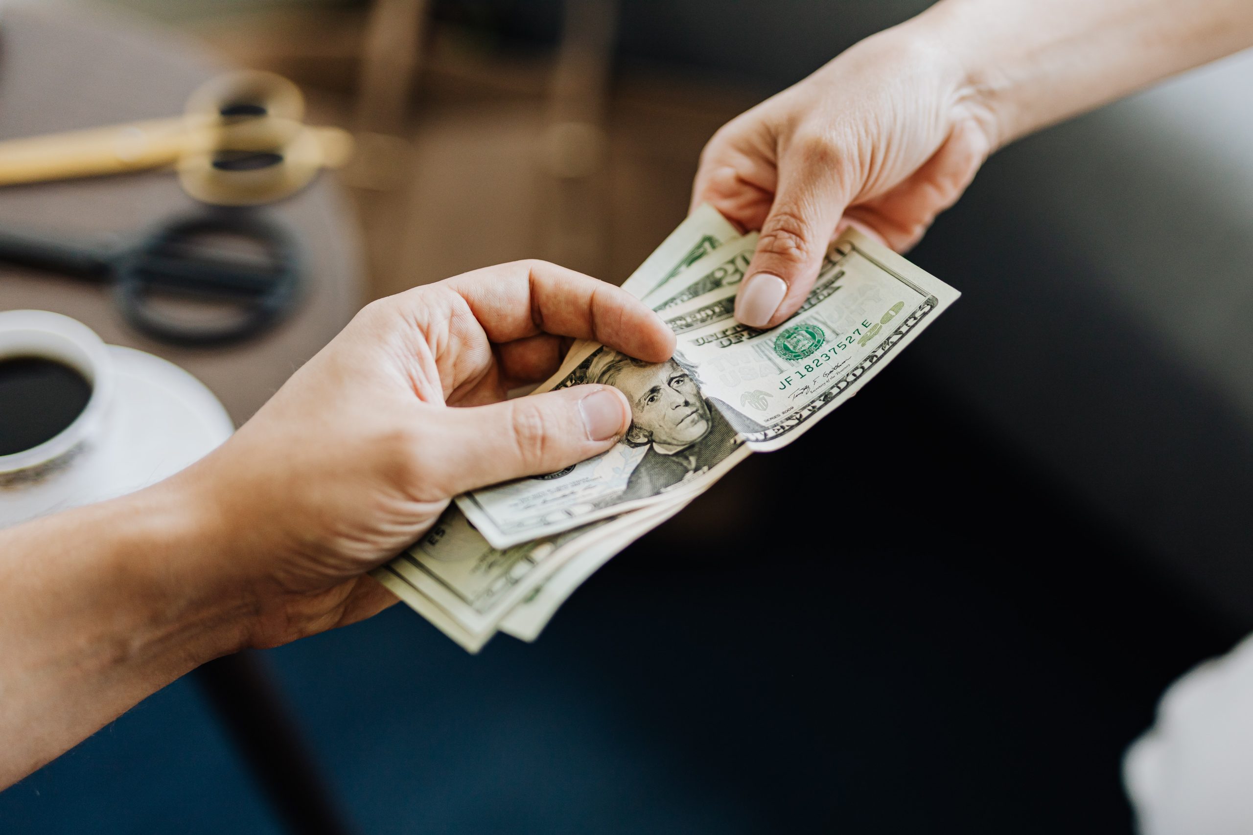 Close-up of two hands exchanging money. One hand, holding multiple US dollar bills, is being handed over to another hand. The background shows a blurred desk with a pair of glasses and a cup of coffee, subtly hinting at financial pitfalls often associated with such transactions. Close-up of two hands exchanging money. One hand, holding multiple US dollar bills, is being handed over to another hand. The background shows a blurred desk with a pair of glasses and a cup of coffee, subtly hinting at financial pitfalls often associated with such transactions.